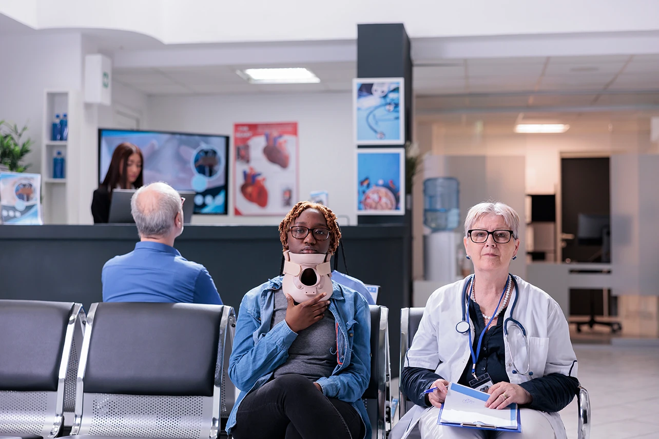 Woman wearing a neck brace sitting in a medical waiting room with a doctor beside her